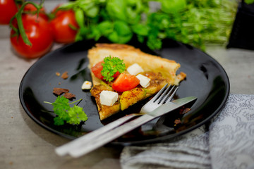 Slice of homemade tart with grilled zucchini, tomatoes and goat's cheese on black plate