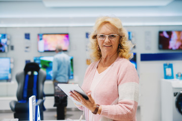 Fototapeta na wymiar Smiling Caucasian senior woman with blonde hair and eyeglasses, dressed casual looking at camera and holding tablet while standing in the tech store.