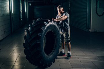 Obraz na płótnie Canvas Side view of muscular attractive caucasian bearded man standing and holding crossfit tyre in underground hallway. Storage units in background.