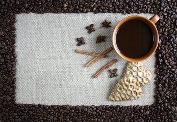 A border of coffee beans, inside on a jute fabric a cup of black coffee, gingerbread hearts, cinnamon and anise