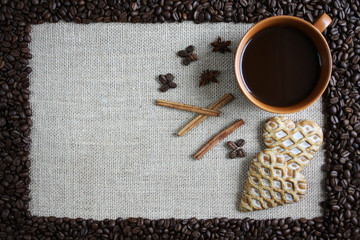 Obramowanie z ziaren kawy, wewnątrz na tkaninie z juty filiżanka czarnej kawy, piernikowe serca, cynamon i anyż