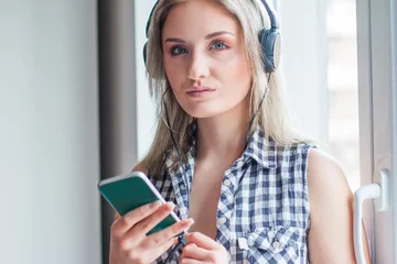 Foto op Plexiglas     Beautiful girl in plaid shirt with headphones listening music indoors and typing on smart phone, blurred background, head and shoulders, youth music concept  © ilijaa
