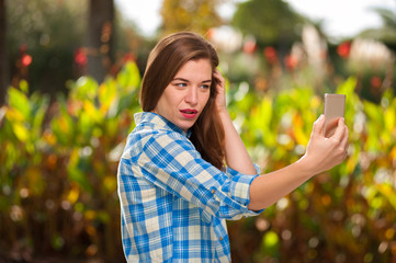 Beautiful young woman taking selfie on a smartphone against the backdrop of a green sunny park on a warm summer day