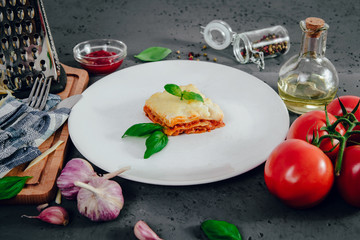 Lasagne on a white plate. On the stone table lies lasagna, spices and food ingredients. The concept of eating and preparing lasagna in tomato sauce.