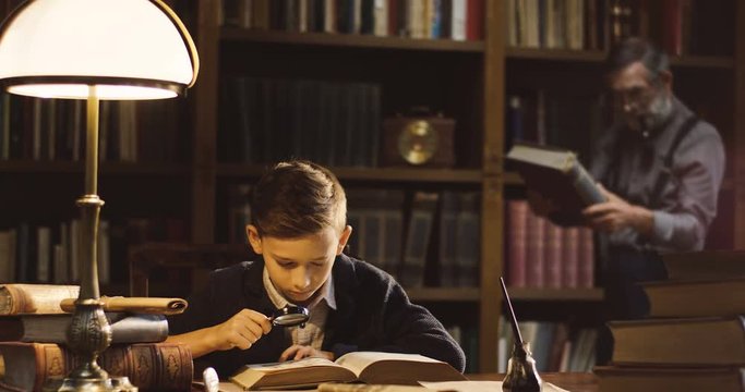 Little cute boy examining pictures in the book with a magnificent glass in hand while his grandpa looking at the book on the background in the ancient library.