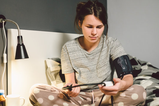 The Topic Of High Blood Pressure Is Hypertension Disease. 22 Year Old Young Caucasian Woman Uses An Automatic Tonometer To Check Pressure At Home In The Bedroom On The Bed In Home Clothes In Evening