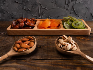 Dates, dried apricots and kiwis in a Compartmental dish and nuts in a wooden spoon on a dark wooden table.