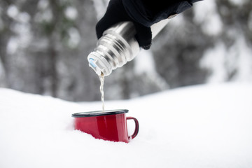 Pouring out tea from a thermos into a cup. Red cup of hot tea on a snow winter background.  