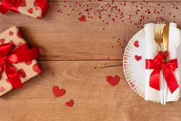 Festive table setting for Valentine's Day with fork, knife, red bow, gifts and hearts on a wooden table. Space for text. Top view