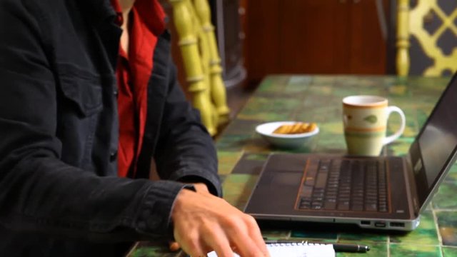 Man at the table is working with laptop. Young man wearing a black jacket and a red shirt is at his laptop, scribbles down notes, tears page apart from the booklet and leaves. Traveling up