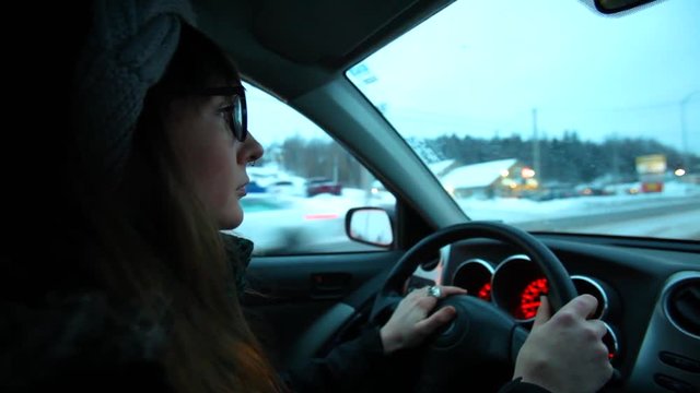 Girl driving at night in the winter. Young woman is driving through her small city in the evening. Blurred lights in the background. Fixed angle scene