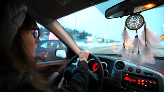 Girl driving at night in the winter. Young woman is waiting at the red light while other cars are passing at her. Blurred lights in the background. Fixed angle scene