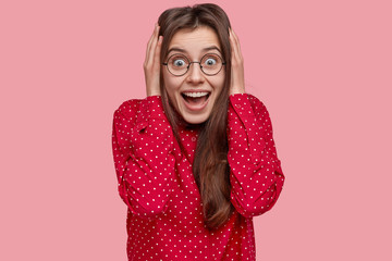 Headshot of amazed brunette girlfriend stares with joyful expression, smiles broadly, shows white teeth, recieves proposal, wears polka dot shirt, isolated over pink background. Emotions concept