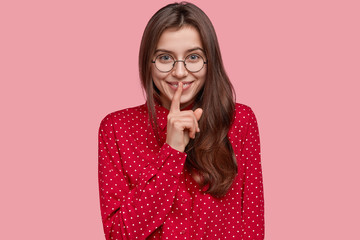 Say no more. Cheerful brunette young lady shows shush gesture with index finger, being glad, gossips with friend, asks not tell her secret, wears red elegant shirt, models over pink studio wall