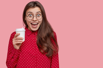 Studio shot of pleased young woman laughs positively, drinks takeaway coffee, has break after lectures, dressed in red clothes, isolated over pink background with copy space for your promotion