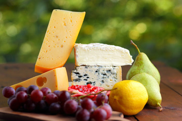 Camembert, brie, hard cheese and grapes on a wooden stand. Pieces of different cheeses, pear and lemon on blurred background. Dorblu, soft cheese and pomegranate on boards. Cheese and fruit