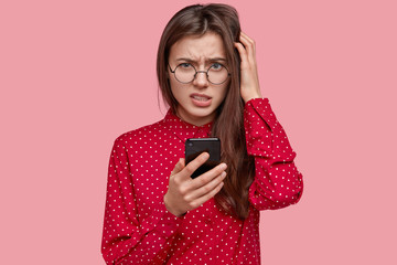 Photo of displeased dark haired woman looks with puzzlement, holds mobile phone, ponders on content...