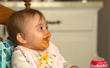 Fototapeta premium Infant laughing while eating dinner with food smeared on her face and down her bib
