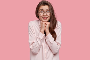 Beautiful young lady keeps hands under chin, has eyes full of happiness, long dark hair, dressed in stylish shirt, wears spectacles, isolated over pink background. Satisfied model feels delighted