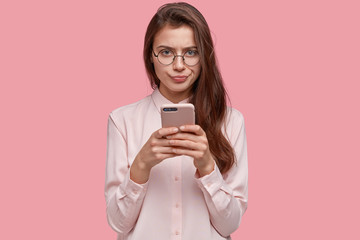 Image of serious girlfriend with displeased facial expression, presses lips, carries modern cell...