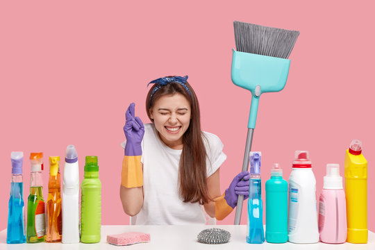 Image Of Wishful Pretty Young Woman Crosses Fingers For Good Luck, Wants To Get Money Reward From Client, Works In Cleaning Service Company, Holds Broom, Uses Multipurpose Chemical Supplies.