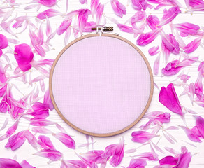 Tools for cross stitch. A hoop for embroidery, canvas and peony petals on white wooden background....