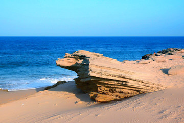 PAISEJA DE PLAYA CON ROCA EN EL CENTRO DESGASTADA POR LAS OLAS Y HORIZONTE DE CIELO AZUL