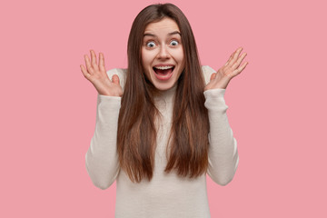 Headshot of attractive excited woman spreads palms near face, has surprised expression, has long hair, dressed casually, isolated over pink studio wall. Happy Caucasian lady feels overjoyed, gestures