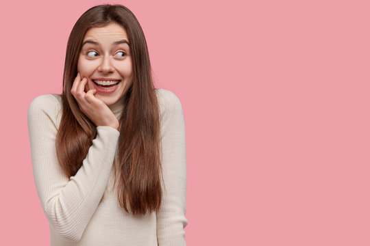 Studio shot of cheerful young European woman keeps hand on cheek, looks positively aside, giggles at something funny, models over pink tudio wall with empty space for your advertising content