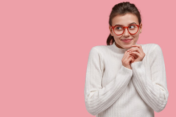 Optimistic dreamy young woman keeps hands together, focused aside with gentle smile, has intention or plan for something grandiose, wears glasses for good vision, isolated over pink studio wall