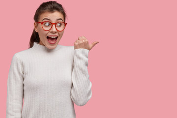 Studio shot of positive happy youngster laughs and points aside, shows something amazing, dressed in casual clothes, isolated over pink studio wall, advertises good with big discounts in shop