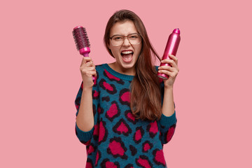 Image of desperate beautiful woman shouts angrily, cant make hairstyle, being angry with hairdresser, carries comb and spray, has long straight hair, models over pink studio wall, yells in annoyance