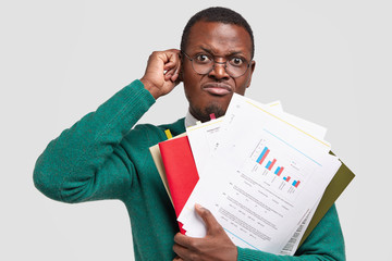 Dissatisfied black man prepares accounting report, holds papers with inforgraphic, wears spectacles...