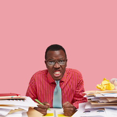 Studio shot of irritated dark skinned man clenches teeth, keeps hands in fists, dressed in formal clothes, writes with pen, looks angrily at camera feels peevish because of much work isolated on pink