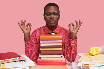 Black relaxed young male manager practices yoga at work place, takes rest for wellness, avoids...