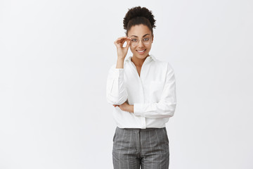 Good-looking stylish and smart female psychologist with dark skin in white-collar shirt and pants, touching glasses and smiling with self-assured expression, gazing at camera