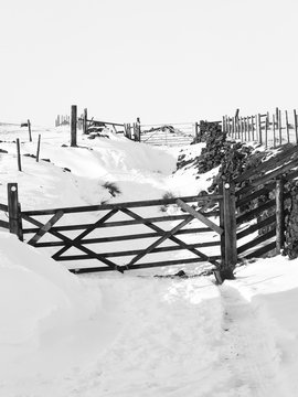 monochrome image of a snow covered country lane with wooden gates and surrounding fields and moors