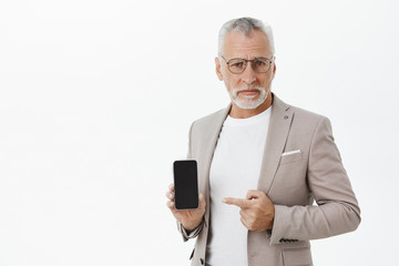 Portrait of unimpressed unsure and hesitant elegant wealthy mature businessman in glasses and suit holding smartphone pointing at device screen and raising eyebrow in dislike over white wall