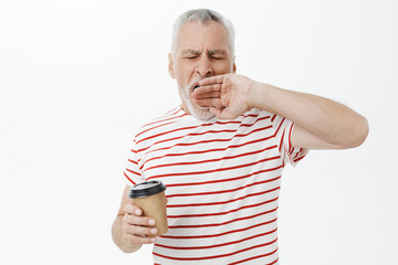Waist-up shot of sleepy and tired grandfather with grey hair yawning with closed eyes holding paper...
