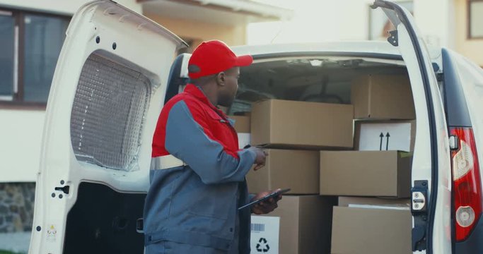 African American young mailman in red costume and cap counting the mail boxes in a van with tablet device in hands. Outdoors.