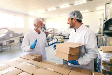 Two happy smiling colleagues talking about work. Older one holding tablet while younger one relocating boxes. Food factory interior.