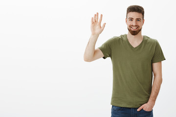 Friendly-looking charming bearded boyfriend feeling shy waving with raised hand in hello or hi gesture, holding hand in pocket casually as meeting new people, greeting over white background © Cookie Studio