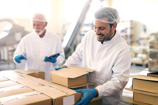 Young Caucasian man relocating boxes with cookies in food factory. In background his boss counting boxes and holding tablet.