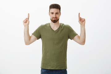 Naklejka premium Guy assured you need check out product, pointing with raised index fingers up at copy space with confident smirk, looking strict and self-assured at camera encouraging try it over white background