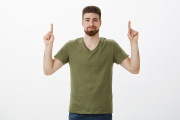 Fototapeta premium Portrait of dreamy delighted and cute bearded boyfriend in love smirking pleased and releaved looking, pointing up with raised hands as finding awesome gift for valentines day against white background