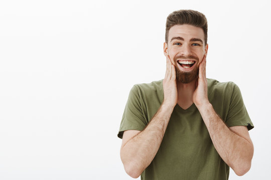 Cheeks hurt from laughing and smiling. Portrait of amused happy upbeat attractive bearded adult male in olive t-shirt touching face and grinning having fun being in great mood over white background