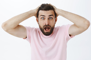 Waist-up shot of shocked man in panic yelling opening mouth combing hair with hands on head staring worried and anxious at camera standing displeased and nervous against gray background