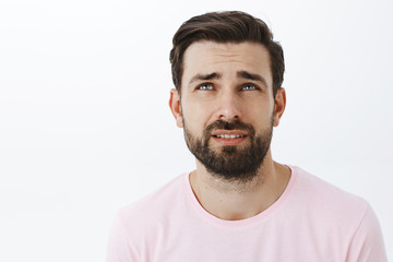 Fototapeta premium Waist-up shot of gloomy and unhappy adult bearded man with blue eyes frowning looking up bothered with regret standign displeased and concerned over gray wall overthinking feeling sad