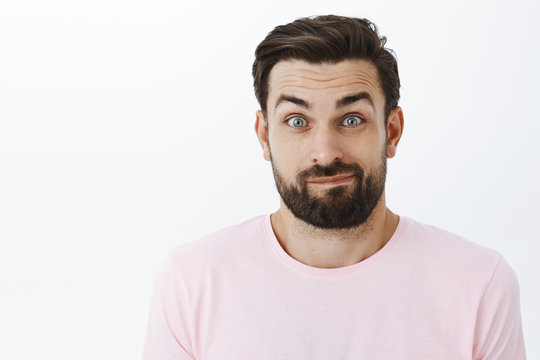 Close-up shot of awkward unsure good-looking caucasian man with beard smirking unsure and silly popping eyes and raising eyebrows clueless posing uncertain and clumsy against gray background