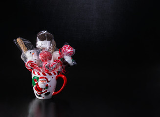 White Christmas mug with Santa Claus picture filled with wrapped cake pops, marshmallows and candy canes on a black background with text space.  Christmas baked gifts.